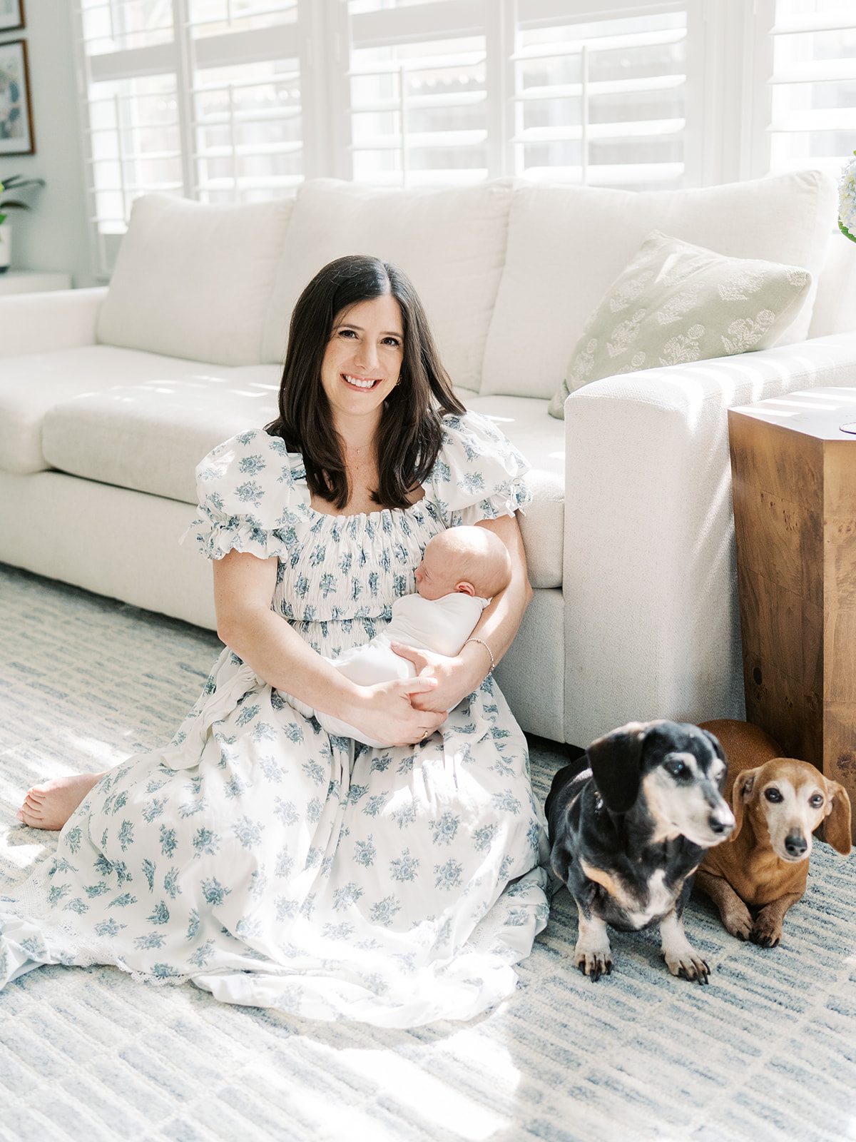 mom holds sleeping newborn while two dogs lay on the floor nearby