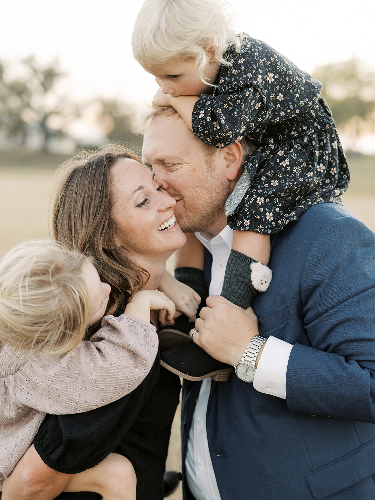 husband kisses wife's cheek while they hold their children 