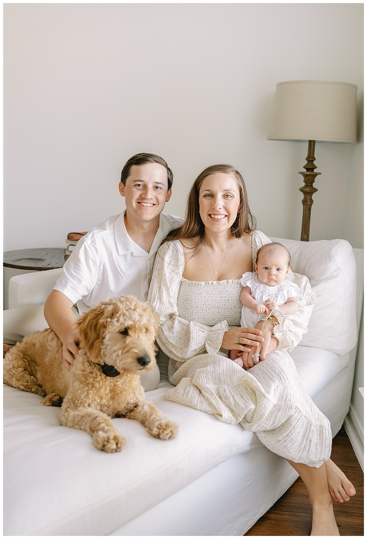 Mom and dad sit on chaise lounge with newborn baby girl and Goldendoodle for their in-home newborn pictures in San Antonio, TX taken by Teressa Jane Photography, a San Antonio Newborn Photographer.